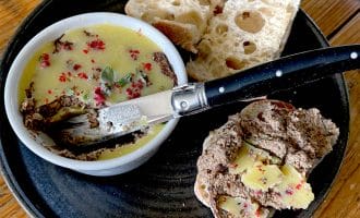 A black plate with a ramekin of pâté topped with herbs and spices, slices of rustic bread, and a knife spread with pâté and butter on a wooden table.