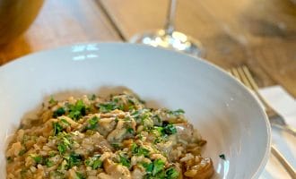 A white bowl of creamy mushroom risotto garnished with chopped parsley sits on a wooden table beside a glass of white wine, with a fork and serviette nearby.