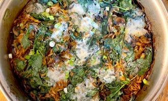 A pot of cooked orzo pasta mixed with spinach, mushrooms, spring onions, and melted cheese, sitting on a wooden table. The ingredients are combined and lightly browned on top.
