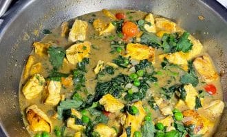 A stainless steel pan filled with a chicken curry dish, featuring chunks of chicken, peas, cherry tomatoes, fresh coriander, and a creamy sauce. A bowl of white rice is visible in the background.