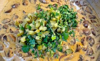 A creamy mushroom stew in a saucepan, topped with chopped gherkins and fresh parsley.