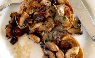 Two slices of toasted bread topped with sautéed mushrooms and herbs on a white plate, with a fork and knife placed beside the food.