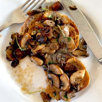 Two slices of toasted bread topped with saut&eacute;ed mushrooms and herbs on a white plate, with a fork and knife placed beside the food.
