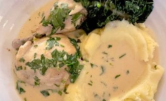 A plate with creamy mashed potatoes, chicken in herb sauce, and a serving of cooked dark leafy greens, all garnished with chopped fresh herbs.