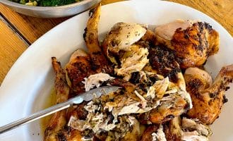 A platter of roasted, carved chicken with metal tongs, served on a wooden table next to a bowl of fresh green salad with broccoli and nuts.