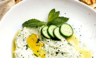 A white plate with creamy tzatziki dip, garnished with olive oil, mint leaves, and cucumber slices. Pieces of flatbread are in the background.