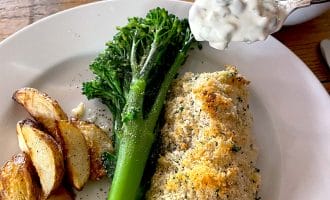 A plate with baked fish topped with breadcrumbs, roasted potato wedges, and steamed tenderstem broccoli. A spoonful of creamy tartare sauce is being served from a small bowl.
