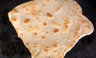 A piece of unevenly shaped flatbread with golden-brown spots cooks on a dark, oily griddle.