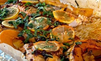 Baked salmon fillet topped with lemon slices and fresh herbs, surrounded by roasted orange and yellow vegetables on a foil-lined baking tray.