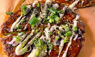Two savoury Japanese pancakes (okonomiyaki) topped with mayonnaise, chopped spring onions, red onions, sesame seeds, and herbs, sitting on brown baking parchment.