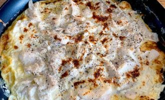A black frying pan on a wooden table contains a cooked, golden-brown omelette topped with melted cheese and black pepper. The omelette appears fluffy with slightly crispy edges.