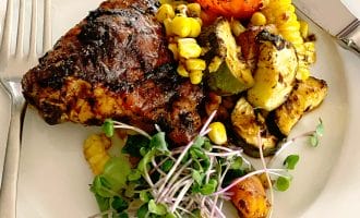 A plate with grilled chicken, roasted vegetables including courgette, tomato, and sweetcorn, garnished with fresh microgreens. A fork and knife are placed beside the food on the white plate.