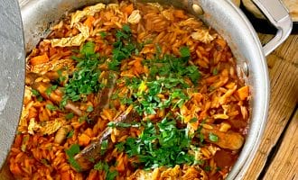 A large saucepan filled with a stew containing sausages, vegetables, orzo pasta, and chopped herbs on top. Flatbread and a glass are placed on the wooden table beside the saucepan.