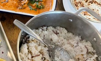 A pot of cooked white rice with cashews and onions sits next to a dish of creamy orange curry garnished with coriander and a plate of round flatbread on a wooden table.