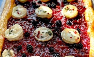 Rectangular puff pastry tart topped with a red jam-like layer, blackberries, and round slices of goats' cheese, garnished with herbs and pink peppercorns. The crust is golden and flaky.