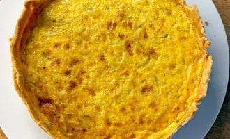 A golden-brown quiche with a flaky pastry sits on a white plate atop a wooden surface. The filling appears smooth and slightly browned on top.