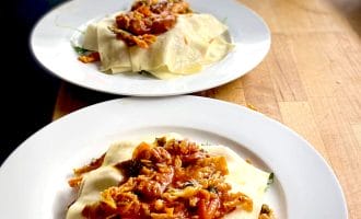 Three white plates sit on a wooden counter, each with homemade lasagne featuring visible pasta sheets and a chunky tomato-based sauce with meat and vegetables, ready to be served.