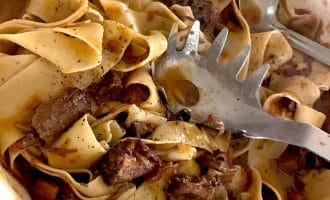 Wide ribbons of pasta tossed with chunks of braised beef in a rich, dark sauce, served in a white dish with a pasta server and metal spoon.