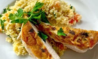 A white plate with grilled chicken breast slices garnished with parsley, served alongside a mound of couscous mixed with chopped vegetables.