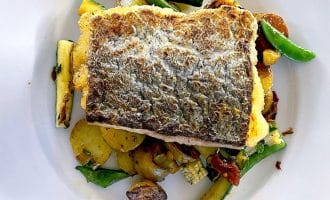 A fillet of fish with crispy skin served on sautéed vegetables, including courgette, potatoes, and green beans, on a white plate.