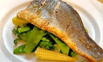 A cooked fish fillet with crispy skin served on a bed of baby corn, broccoli, and mangetout, presented on a white plate.