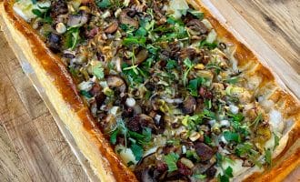 A rectangular puff pastry tart topped with sliced mushrooms, fresh herbs, and onions sits on baking parchment on a wooden surface. A knife is visible nearby.