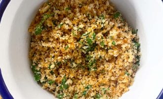 A white bowl with a blue rim filled with golden-brown seasoned breadcrumbs mixed with chopped green herbs, sitting on a wooden surface.