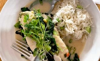 A white bowl with steamed white fish topped with fresh herbs and greens in a light sauce, served alongside a portion of white rice with spring onions. A fork and spoon rest on the edge of the bowl.