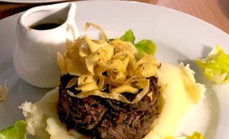 A plated dish featuring shredded beef topped with crispy fried onions, surrounded by creamy mashed potato and garnished with green leaves, with a small jug of brown sauce on the side.