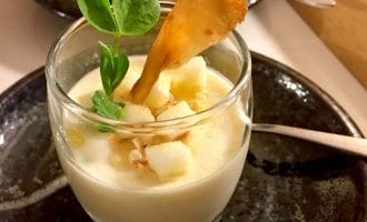A glass dessert dish filled with creamy panna cotta, topped with diced fruit, a crunchy garnish, and a small green sprig, placed on a dark plate with a spoon beside it.