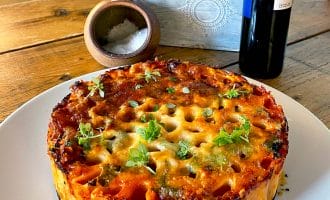 A baked pasta pie with a golden, cheesy top and herbs is served on a white plate. There’s a wooden table, a salt pot, a bottle, and a planter with greens in the background.