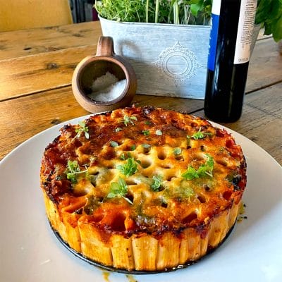 A baked pasta pie with a golden, cheesy top and herbs is served on a white plate. There&rsquo;s a wooden table, a salt pot, a bottle, and a planter with greens in the background.
