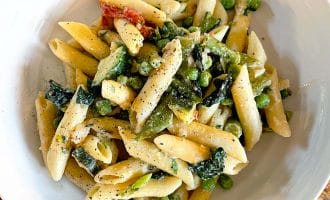 A white bowl filled with penne pasta mixed with green peas, leafy greens, and a creamy sauce, topped with cracked black pepper, sits on a wooden table.