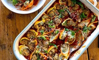 A baking dish filled with stuffed pasta shells topped with tomato sauce and herbs, with a serving spoon and a bowl of mixed green salad beside it on a wooden table.