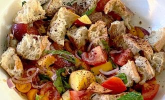 A bowl of panzanella salad with chunks of rustic bread, tomatoes, yellow cherry tomatoes, peach slices, red onion, fresh basil, and ground black pepper.