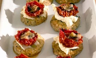 A white rectangular platter holds nine savoury appetisers made with a round biscuit base, topped with creamy cheese, roasted tomato halves, herbs, and small mushroom slices.