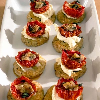 A white rectangular platter holds nine savoury appetisers made with a round biscuit base, topped with creamy cheese, roasted tomato halves, herbs, and small mushroom slices.