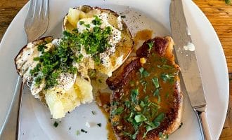 A white plate with a baked potato topped with soured cream, chopped chives, and pepper, next to a pork chop with brown sauce and herbs. A fork and knife are placed on the plate. The table is wooden.
