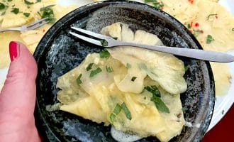 A hand holds a dark ceramic bowl with thinly sliced pineapple, garnished with fresh herbs. In the background, more pineapple slices with herbs and red chilli are arranged on a white plate. A small fork rests in the bowl.
