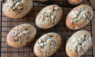 Oval-shaped baked goods topped with chopped nuts are cooling on a black wire rack placed on a wooden surface. The golden-brown pastries appear fluffy and freshly baked.