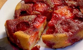 A close-up of a plum upside-down cake on a white plate, with a slice being lifted out. The cake has caramelised plums on top and a golden, moist-looking base. The background is a wooden surface.