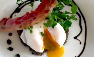A poached egg with a runny yolk sits on a dark patty, garnished with microgreens and chives, topped with a crispy rasher of bacon, and surrounded by a swirl of dark sauce on a white plate.