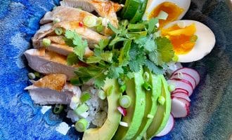 A blue bowl filled with rice, sliced grilled chicken, avocado, soft-boiled egg, cucumber, radish, coriander, and spring onions, arranged neatly for a colourful and fresh meal.