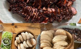A platter of sliced, glazed pork fillet, a bamboo steamer filled with fluffy bao buns, and plates of spring onions, cucumber, and hoisin sauce arranged on a wooden table.