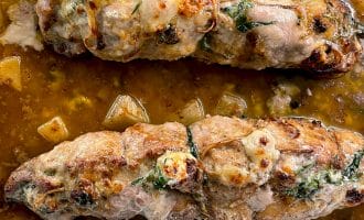 Two stuffed pork fillets tied with string are baked in a brown sauce with visible herbs and chunks of vegetables in a rectangular roasting tin.