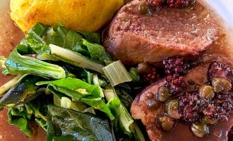 A plate with two slices of beef topped with capers and berries in sauce, sautéed leafy greens, and a round serving of cornbread or a baked yellow side.