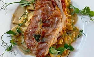 A white plate with pasta topped with a slice of prosciutto-wrapped meat, creamy sauce, and garnished with fresh pea shoots.