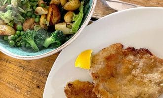 A white plate with two breaded cutlets and a lemon wedge, beside a bowl of roast potatoes, broccoli, peas, and greens on a wooden table.