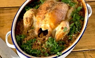 A whole roast chicken sits atop a bed of seasoned grains and herbs in an oval casserole dish, garnished with chopped parsley, on a wooden table beside a carving knife.