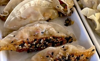 A close-up of pan-fried dumplings topped with white and black sesame seeds, arranged in a white rectangular dish alongside steamed dumplings.